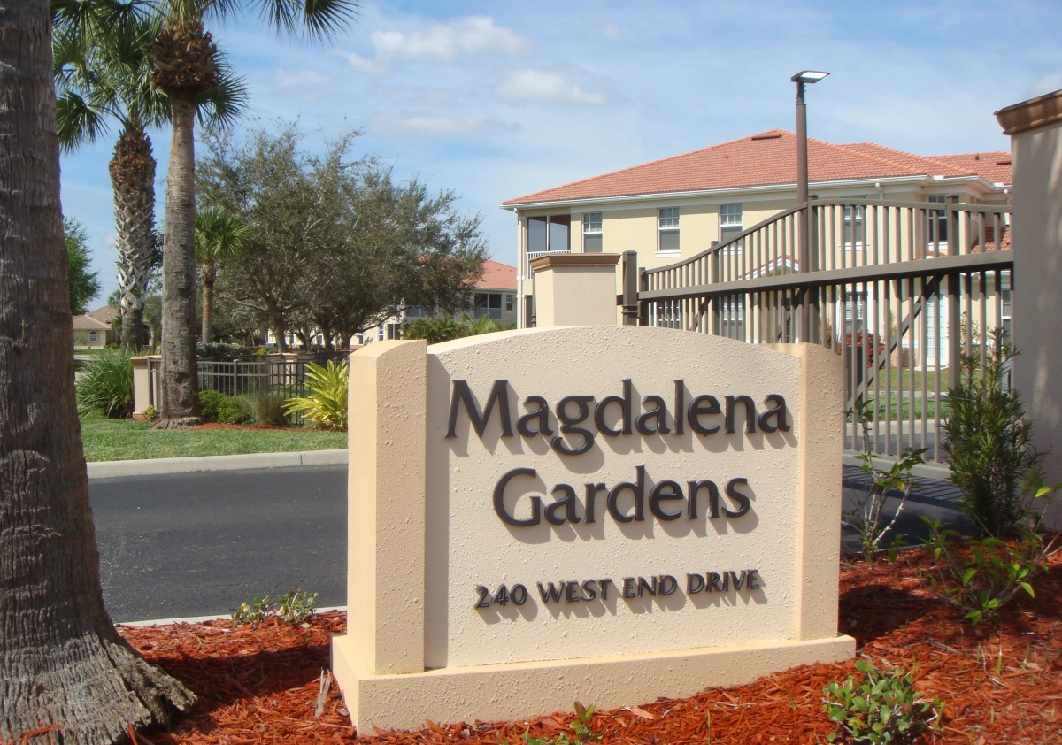 Photo showing a road sign structure that reads "Magdalena Gardens, 240 West End Drive" In the background there is a 2 story building and behind the sign is an access control gate. There is also a living oak tree and several palm trees in the photo.